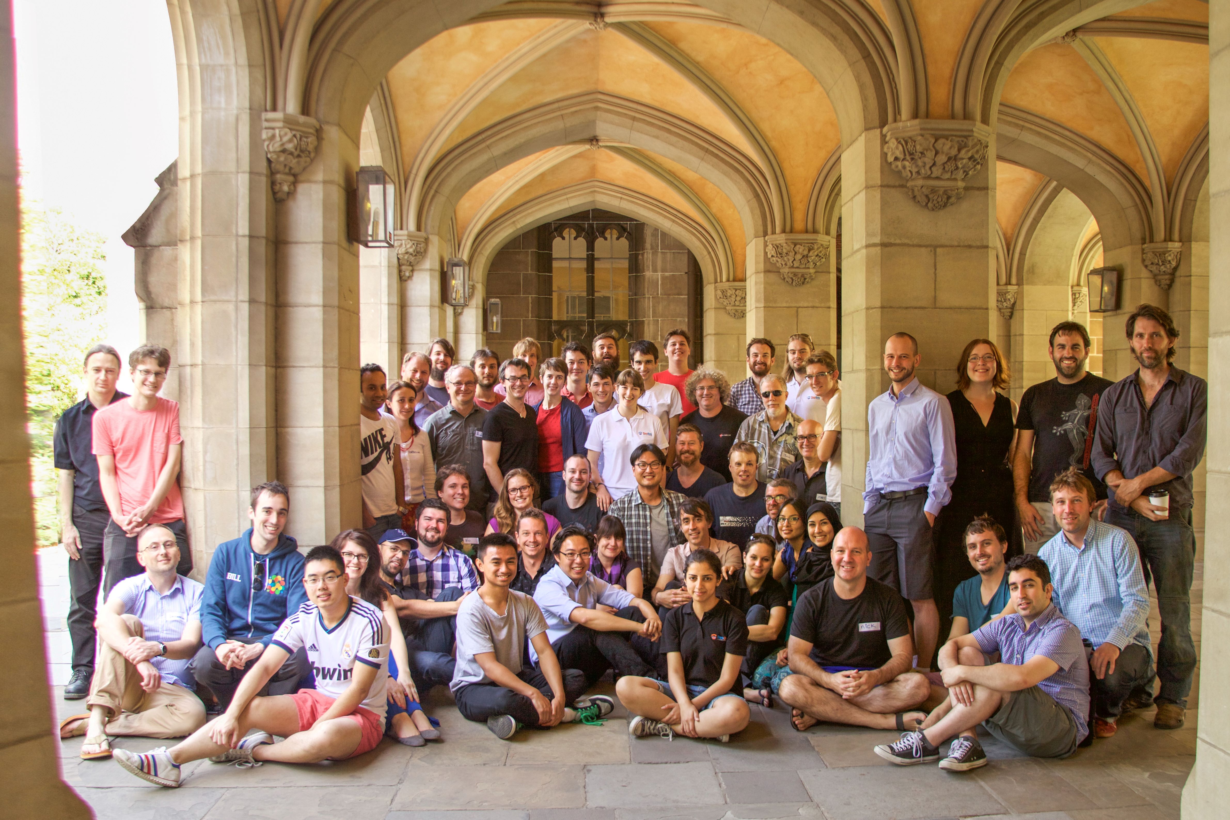 Image of a group of 53 adults standing and kneeling and sitting on a concrete floor surrounded by ornate pillars under a university cloister